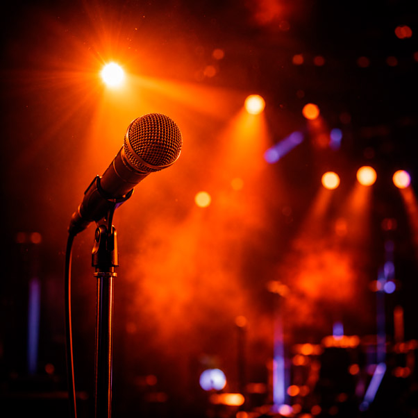 Fotografía de backstage de un concierto o show en vivo: en primer plano, un micrófono profesional sobre pie cromado bajo un haz de luz cálida naranja intensa que genera un halo luminoso y un sutil destello de lente; fondo muy desenfocado con siluetas abstractas de luces de escenario tipo PAR LED en tonos naranja y ámbar, con un contraste puntual en azul/morado oscuro, evocando un concierto en vivo sin mostrar público ni artistas reconocibles; partículas de humo escénico iluminadas flotando en el aire dando textura volumétrica a la luz; composición vertical o cuadrada con bokeh grande y cálido; paleta dominante negro profundo de fondo, naranja y ámbar como colores protagonistas, con toques fríos de azul/violeta para contraste; estilo de fotografía de evento en vivo de alto dramatismo, sensación de energía y espectáculo, sin rostros ni logotipos.