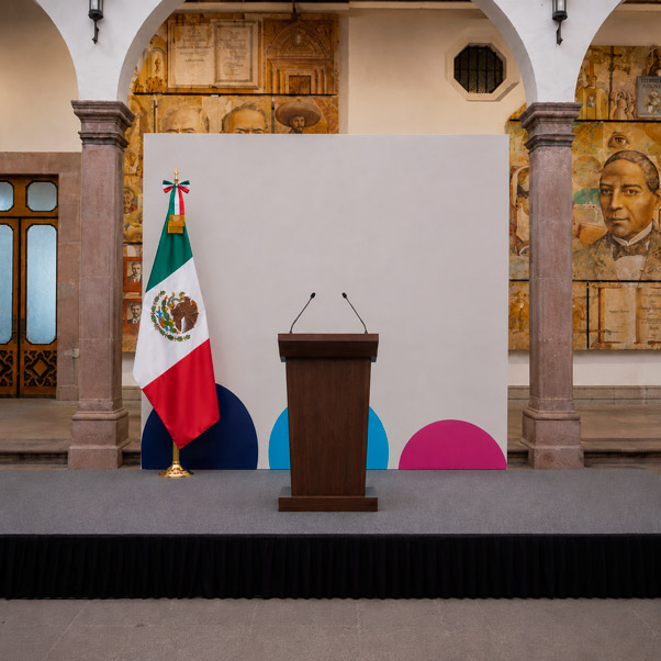 Fotografía institucional de un evento gubernamental en un recinto formal: vista de un templete elevado con atril central de madera oscura y un fondo institucional liso en tono crema con un elemento gráfico geométrico sutil en naranja a modo de franja lateral, sin escudos, banderas ni emblemas oficiales reales; banderas genéricas abstractas, sin diseño de país real, en soportes a ambos lados del atril en tonos blanco con un acento naranja en el asta; el recinto tiene techos altos con columnas clásicas simplificadas que transmiten formalidad institucional; iluminación uniforme cálida tipo evento oficial con un ligero acento de luz naranja rasante sobre el atril; composición horizontal con simetría central marcada y ángulo frontal ligeramente elevado; paleta de crema, blanco, madera oscura y acentos naranja controlados; estilo de fotografía institucional y protocolar, seria pero cálida, sin personas ni emblemas oficiales reconocibles.